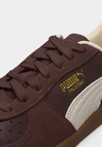 Gros plan sur une basket Puma Palermo marron avec une texture en daim, une bande latérale blanche, des lacets marron et le logo Puma Palermo doré sur le côté.