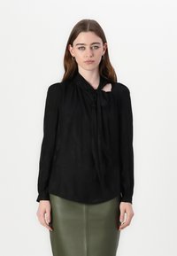 Sort bluse med lange ærmer, sløyfe detalj ved halslinjen, subtil blomsterstruktur, kombinert med et tettsittende olivengrønt skjørt.