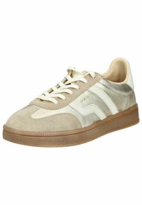 Zapatilla baja en beige y dorado metálico con rayas blancas, cordones blancos y suela de goma marrón texturizada.