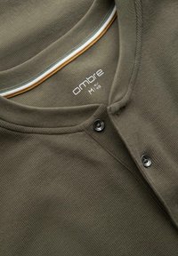 Närbild av en olivgrön strukturerad henley-skjorta med två svarta knappar och randig invändig halsringning.