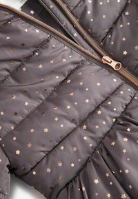 Gros plan sur un tissu de veste matelassée marron avec un motif étoile métallique en or rose et une fermeture éclair en or rose.
