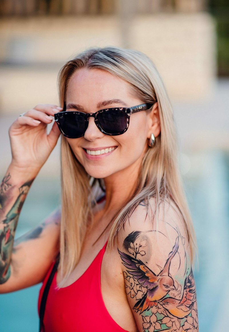 Černé sluneční brýle s mramorovým rámem, které nosí osoba s dlouhými blond vlasy. Mají živé tetování na paži a nosí červené plavky.