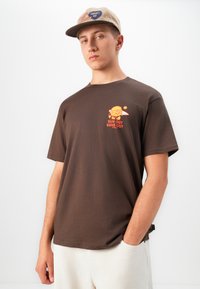 Ung mand iført en brun "Sun Out Buns Out" t-shirt og en beige Carhartt-kasket, med den ene hånd i lommen foran en enkel hvid baggrund.