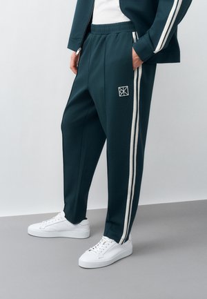 Osoba ubrana w ciemnozielone spodnie dresowe z białymi paskami po bokach i logo, do tego białe trampki i pasująca kurtka.