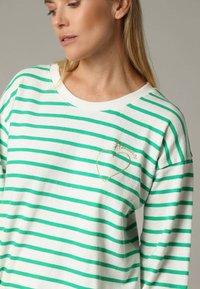 Sweatshirt rayé vert et blanc avec un col rond, texture de tissu douce et un détail en forme de cœur doré sur la poitrine.