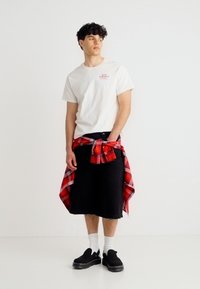 Νέος άνδρας φορώντας λευκό T-shirt, μαύρα σορτς μέχρι το γόνατο, μαύρα παπούτσια με λευκές κάλτσες και κόκκινο πουκάμισο καρό δεμένο γύρω από τη μέση, όρθιος.