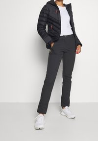 Mujer vistiendo una chaqueta acolchada negra, camiseta blanca, pantalones negros y zapatillas deportivas blancas, de pie contra un fondo blanco sencillo.