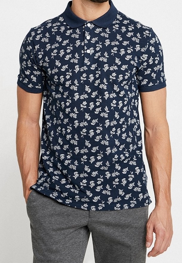 Homme portant un polo à manches courtes bleu marine à motif floral blanc et un pantalon gris, se tenant avec une main dans la poche.
