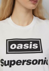 Weißes Baumwoll-T-Shirt mit fettem schwarzen Text, der "oasis" in einer rechteckigen Box und "Supersonic" darunter liest, Rundhalsausschnitt, glatte Textur.