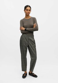 Haut à manches longues marron, pantalon à carreaux gris avec coupe ajustée, et chaussures noires avec une lanière. Le mannequin se tient les bras croisés sur un fond uni.