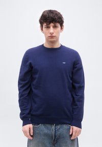 Νέος άνδρας που κοιτάζει προς τα εμπρός, φοράει ένα μπλε σκούρο πουλόβερ Levi's με μακριά μανίκια και ανοιχτό μπλε τζιν, μπροστά σε ένα απλό ανοιχτό γκρι φόντο.