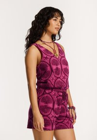 Fukszia ujjatlan overall kagyló mintával, rugalmas derékkal és texturált anyaggal. Kiegészítők között szerepel egy színes gyöngyökkel díszített nyaklánc.