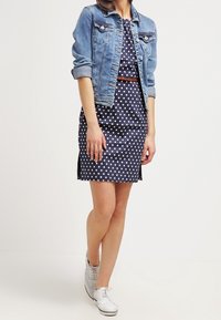 Blå denimjacka över en klänning med prikkar, med figursydd midja och sidofalsor. Vita sneakers fullbordar outfiten, med ett brunt bälte som accent.