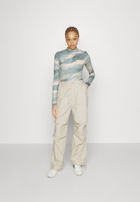 Dug nadstavljeni top s plavim i bež valovitim uzorkom, u kombinaciji s labavim bež hlačama. Outfit uključuje bijele tenisice i minimalistički dizajn.