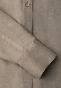 Poignet de chemise beige à manches longues avec une bande ton sur ton verticale et un petit bouton rond sur la patte de boutonnage au-dessus.