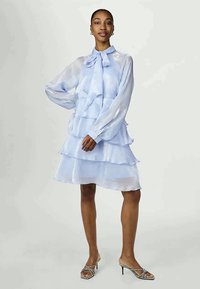 Γυναίκα που στέκεται φορώντας ένα ανοιχτό μπλε στρώμα από σιφόν φόρεμα με μακριά μανίκια και κολάρο με φιόγκο, συνδυασμένο με ανοιχτές κουλούρες με τακούνι.