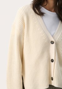 Krémová cardigan s vaflovou pleteninou, výstrihom do V a piatimi gombíkmi. Obsahuje spadnuté ramená a voľný strih.
