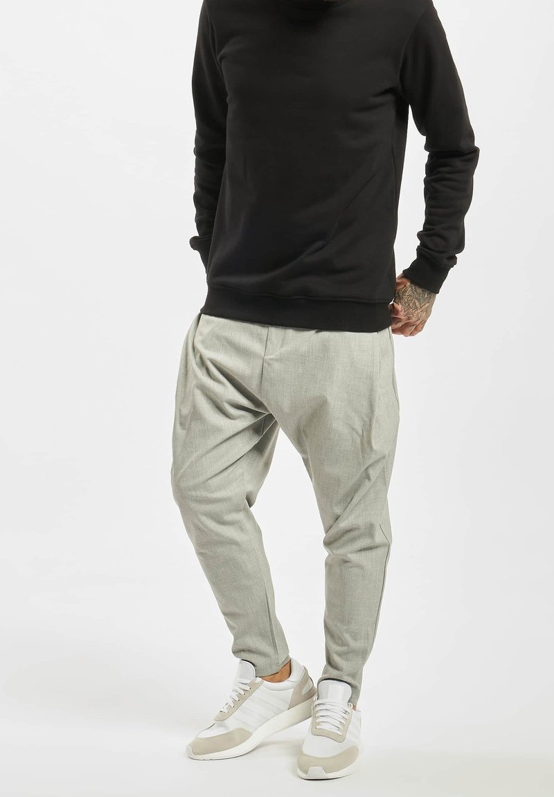 Homme portant un pantalon ample gris clair, des baskets blanches et beiges, et un haut noir à manches longues, debout devant un fond uni.