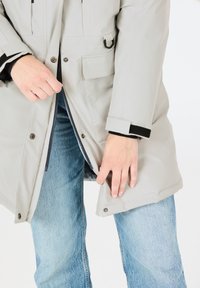 Hellgrauer Parka mit einem Frontreißverschluss und Druckknöpfen, mehreren Taschen und schwarzen Akzenten an den Ärmeln, getragen über blauen Jeans.