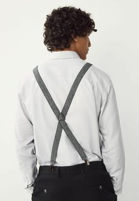Man med lockigt hår som bär en vit långärmad skjorta och svarta byxor, sedd bakifrån, med grå hängslen som korsar sig på ryggen.