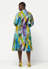 Vestido colorido y estampado que presenta una mezcla de diseños abstractos en azul, amarillo, púrpura y verde. Manga larga y falda acampanada de longitud media.