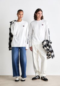 Witte longsleeve shirts met logo patches, gecombineerd met wijdvallende denim en witte broeken. Zwart-wit geruit shirt over één schouder gedrapeerd.