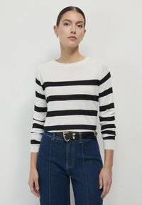Frau, die einen weißen Pullover mit schwarzen horizontalen Streifen, blaue hochtaillierte Jeans und einen schwarzen Gürtel mit einer goldenem Schnalle trägt.