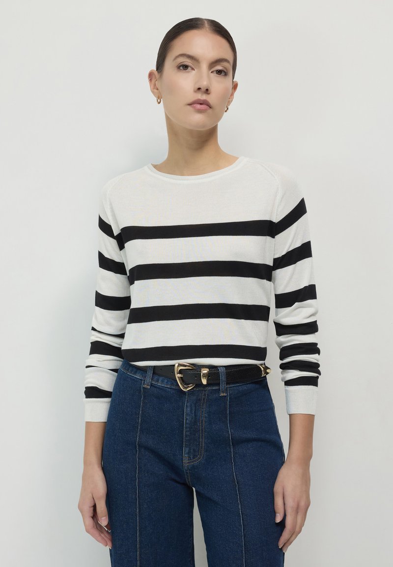 Frau, die einen weißen Pullover mit schwarzen horizontalen Streifen, blaue hochtaillierte Jeans und einen schwarzen Gürtel mit einer goldenem Schnalle trägt.