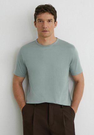 Muž s krátkymi hnedými vlasmi, oblečený v svetlomodrej košeli s krátkym rukávom a tmavohnedých nohaviciach, stojaci s rukami v vreckách pri bielej stene.