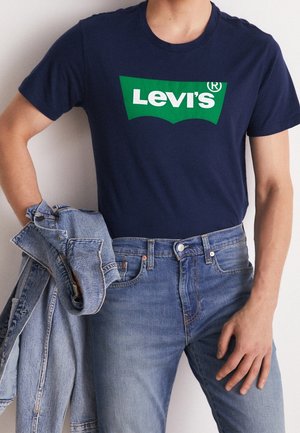 Person iført en marineblå Levi's t-shirt og lyseblå jeans, der holder en sammenfoldet lys denimjakke i den ene hånd.
