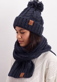 Navy knitted beanie with a pom-pom and ribbed band, paired with a matching scarf. Both feature a textured pattern and leather accents.