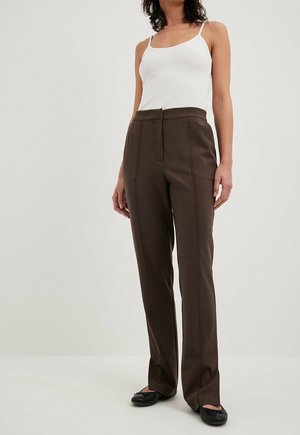 Femme portant un haut blanc sans manches, un pantalon marron foncé taille haute à jambes droites et des ballerines noires, debout devant un fond neutre.