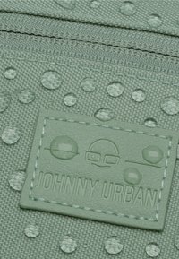 Zelená texturovaná látka s kapkami vody a přišitým štítkem, na kterém je nápis "JOHNNY URBAN." Povrch vykazuje tkaný vzor.