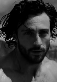 Un primo piano in bianco e nero di un uomo con capelli folti e mossi, una mascella ben definita e la pelle bagnata che scintilla alla luce.