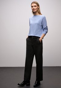 Femme debout les mains dans les poches, portant un pull bleu clair, un pantalon large foncé et des bottines noires, sur un fond uni.