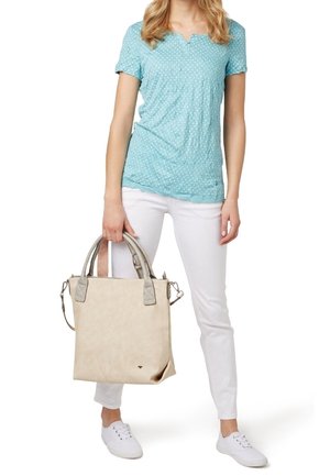 Bolso tote texturizado en tono beige con asas grises, de forma cuadrada y con una correa adicional para el hombro. Se lleva con una blusa de lunares azul clara y pantalones blancos.