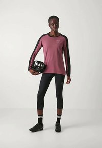 Hosszú ujjú bordó sporttop fekete részletekkel, fekete capri leggings és fekete kerékpáros cipő. A modell egy fekete sisakot tart.