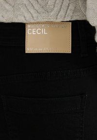 Černé džíny s béžovou koženou nášivkou s embosovaným textem "Cecil" a souřadnicemi. Obsahuje detail zadní kapsy.