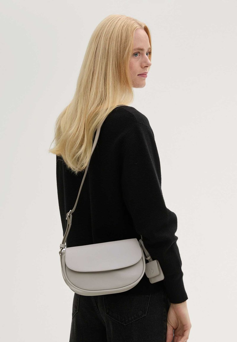 Blonde Frau in schwarzem Pullover trägt eine hellgraue Umhängetasche mit Klappe und kleinem angebrachtem Täschchen, Blick nach rechts vor einfachem Hintergrund.