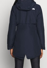 Granatowa kurtka puchowa z kapturem, z wciągniętą talią i elastycznymi mankietami. Tył kurtki zdobi klapa z detalami w postaci przeszyć.