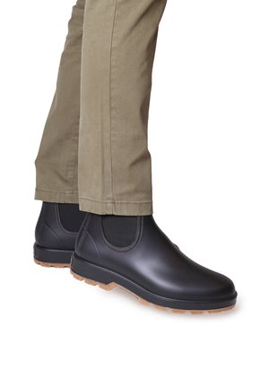 Botas de tobillo impermeables negras con paneles laterales elásticos y suelas de goma, combinadas con pantalones caqui claros. Textura suave y diseño elegante.