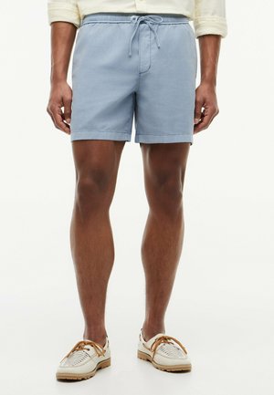 Man draagt lichtblauwe short met aantrekkoord, beige overhemd met lange mouwen opgerold bij de mouwen en witte casual schoenen met bruine veters.