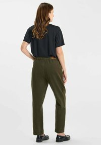 Vrouw met golvend bruin haar staat met haar rug naar ons gekeerd, draagt een zwart T-shirt met korte mouwen, olijfgroene broek en zwarte instapschoenen op een witte achtergrond.