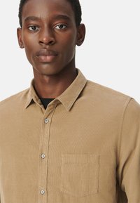 Karmelowa koszula zapinana na guziki z kołnierzykiem, z pojedynczą kieszenią na piersi i subtelnym pionowym prążkowaniem. Gładka tekstura z akcentami guzika.