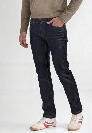Hombre con vaqueros ajustados azul oscuro, camiseta de manga larga beige y zapatillas blancas con rayas azules y rojas, de pie con una mano en el bolsillo.