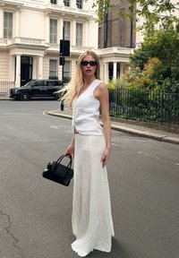 Valge kootud topp V-kaelusega ja tekstuurse viimistlusega, koos pika valge tekstuurse seelikuga; hoiab käes väikest musta käekotti.