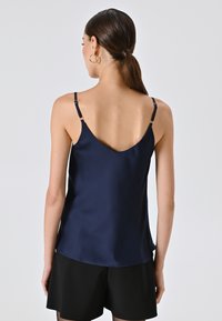 Donna con coda di cavallo scura che indossa una canottiera di raso blu navy con spalline sottili e pantaloncini neri, vista da dietro su uno sfondo semplice.