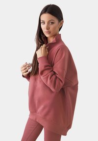 Felpa over-sized con zip in tessuto rosa polveroso, caratterizzata da un colletto alto, polsini a coste e dettagli nelle cuciture laterali. Vestibilità comoda e rilassata.