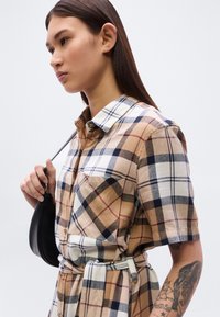 Καφέ πουκάμισο με καρό μοτίβο, κοντά μανίκια και γιακά με κουμπιά, με μπεζ και ναυτικό μπλε λεπτομέρειες. Περιλαμβάνει μικρή τσέπη στο στήθος και ζώνη στη μέση.