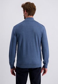 Uomo con capelli castani che indossa un maglione blu a maniche lunghe e pantaloni neri, in piedi con la schiena rivolta verso uno sfondo chiaro e uniforme.